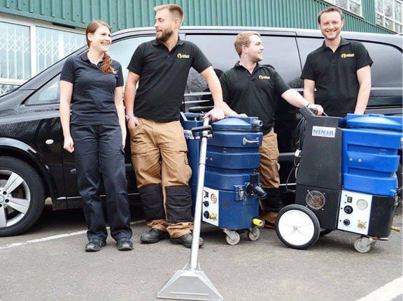 This is a photo of Penge Carpet Cleaning carpet cleaners (three men and one woman) standing in fromt of their black van, with two steam cleaning carpet machines next to them